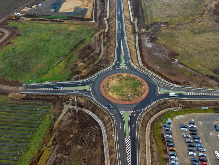 A slight bird's eye view of a road under construction. In the foreground you can see a newly constructed roundabout, in the background a bridge leads over a railway line.