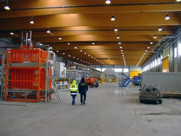 Photo: well-lit, brand-new factory, machines and containers on the sides; in the middle two workers viewed from the back walking away and wearing PORR-branded working clothes and a safety jacket; on the right side, a car covered in dust and gateways; at the top, ceiling with lamps