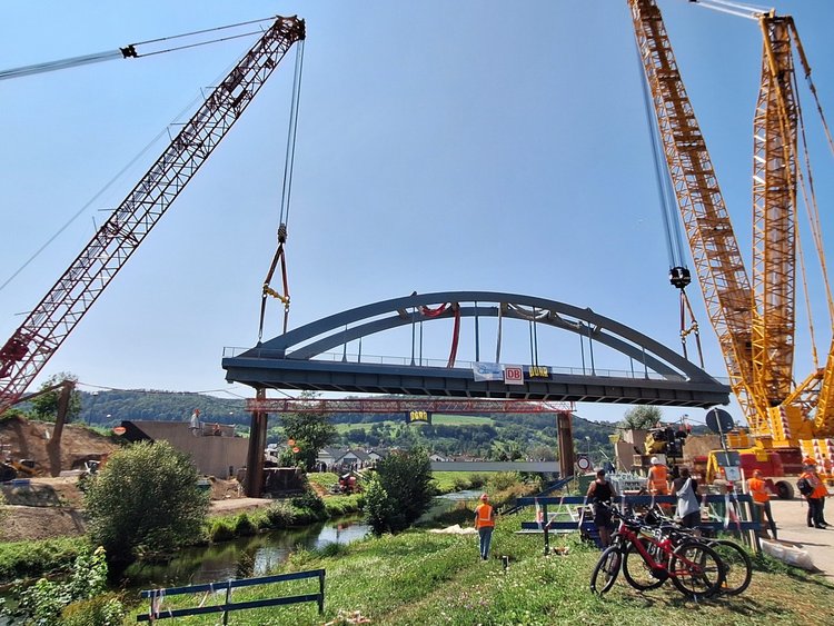 Two large cranes lift a steel bridge over a river.