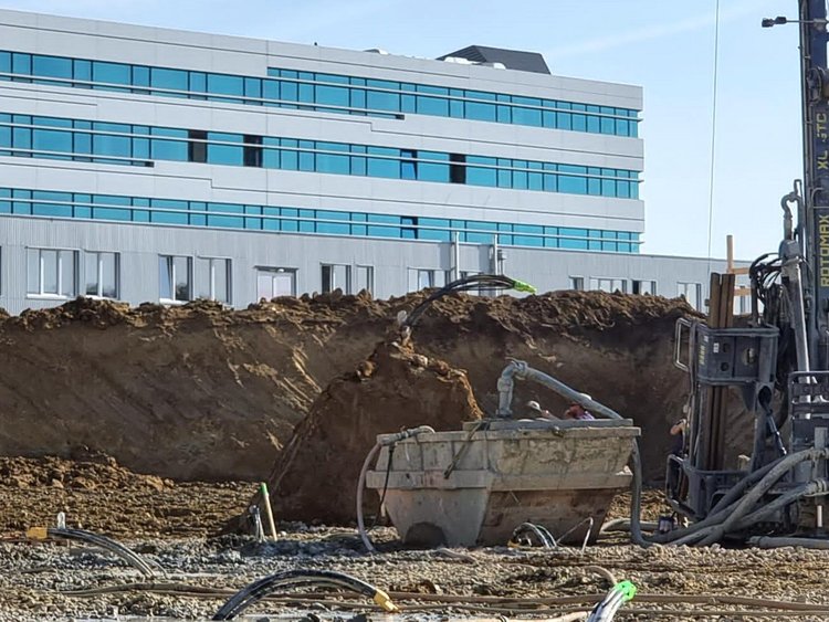 In the foreground, a construction site with a geothermal drilling rig and a skip, with pipes protruding from the ground. In the background is the existing FH Campus Wien building.