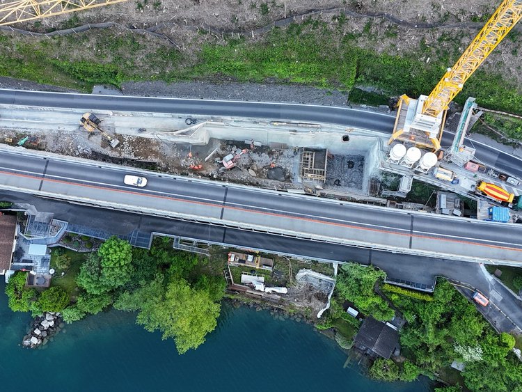 Photo: Aerial view of the construction site with large crane. The road is used by cars and is located between the lake and the steep rock face.