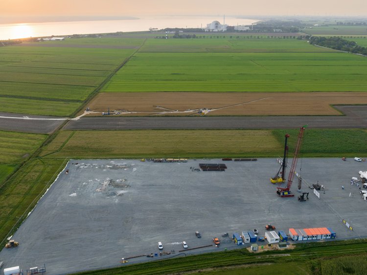Blick aus der Luft auf viele grüne Felder neben einem großen Fluss. Inmitten der Felder ist ein großes graues Baufeld erkennbar, auf dem erste Baucontainer, eine Ramme und ein Schlitzwandgreifer stehen. 