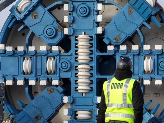 A close-up of a blue tunnel boring head. A member of staff wearing a yellow high-visibility vest is standing in front of it with his back to the camera. 