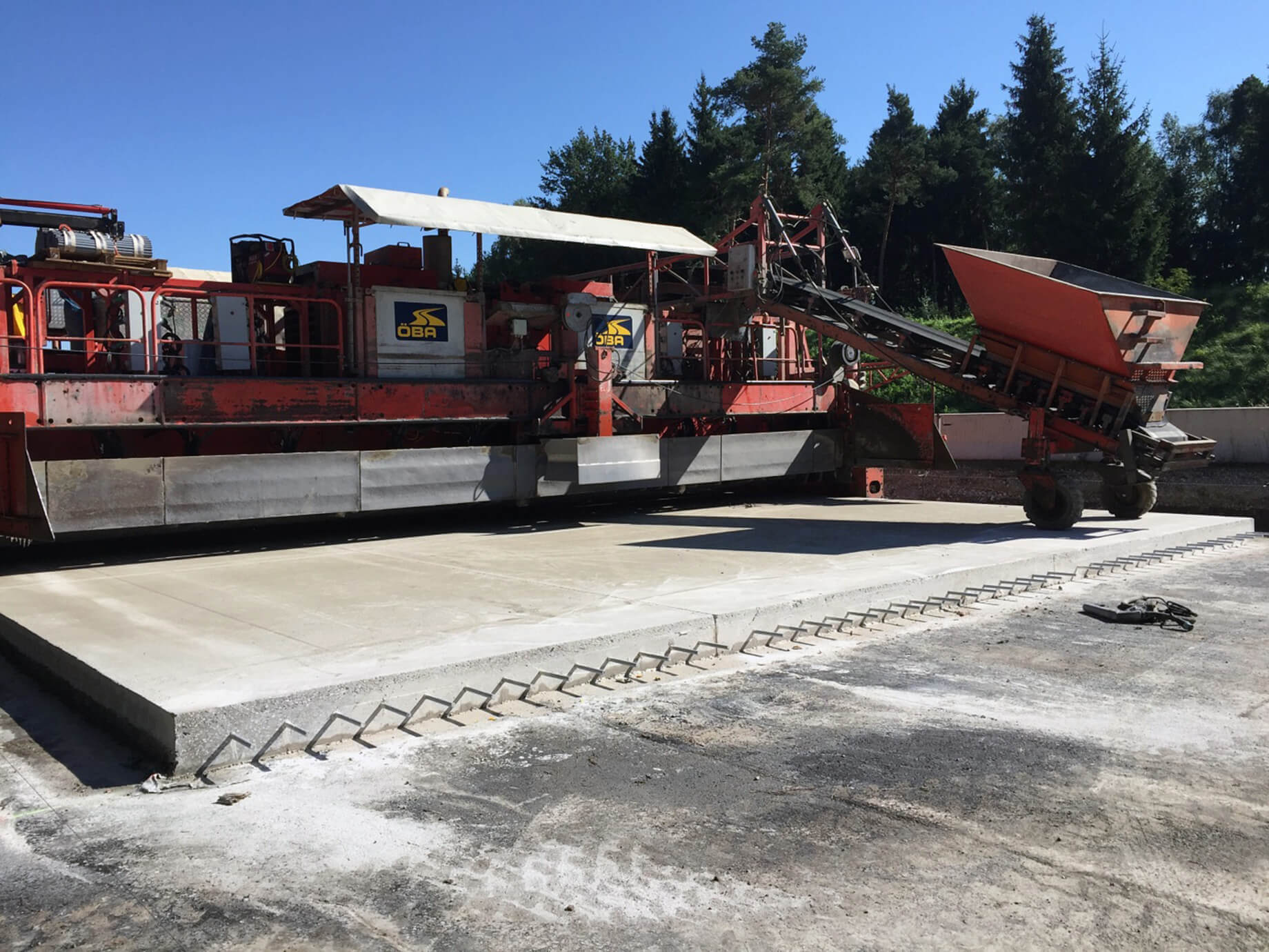 Photo: ÖBA’s bottom concrete paver with a prepared bottom concrete slab with dowels protruding from the longitudinal side, trees on the top right