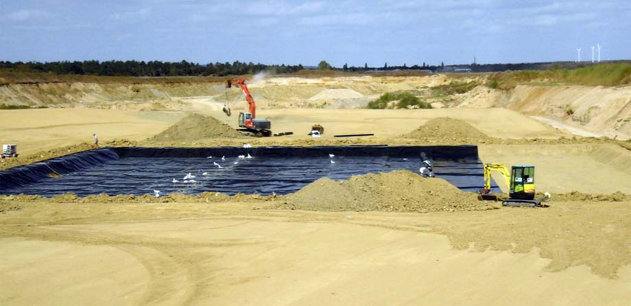Photo: Spacious, level surface of pale brown sand; in the middle, an area covered in black foil, construction vehicles transporting soil material.