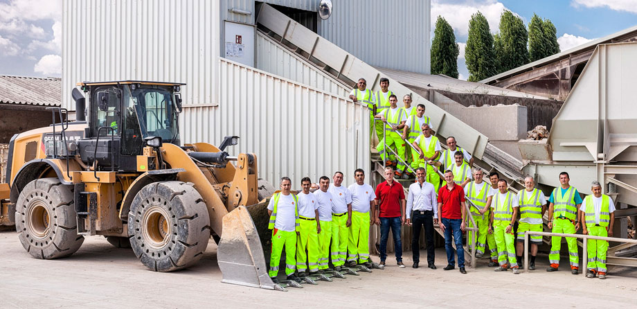 Photo: Group of around 20 men, some of them in work clothes, posing in front of an industrial plant and a wheel loader