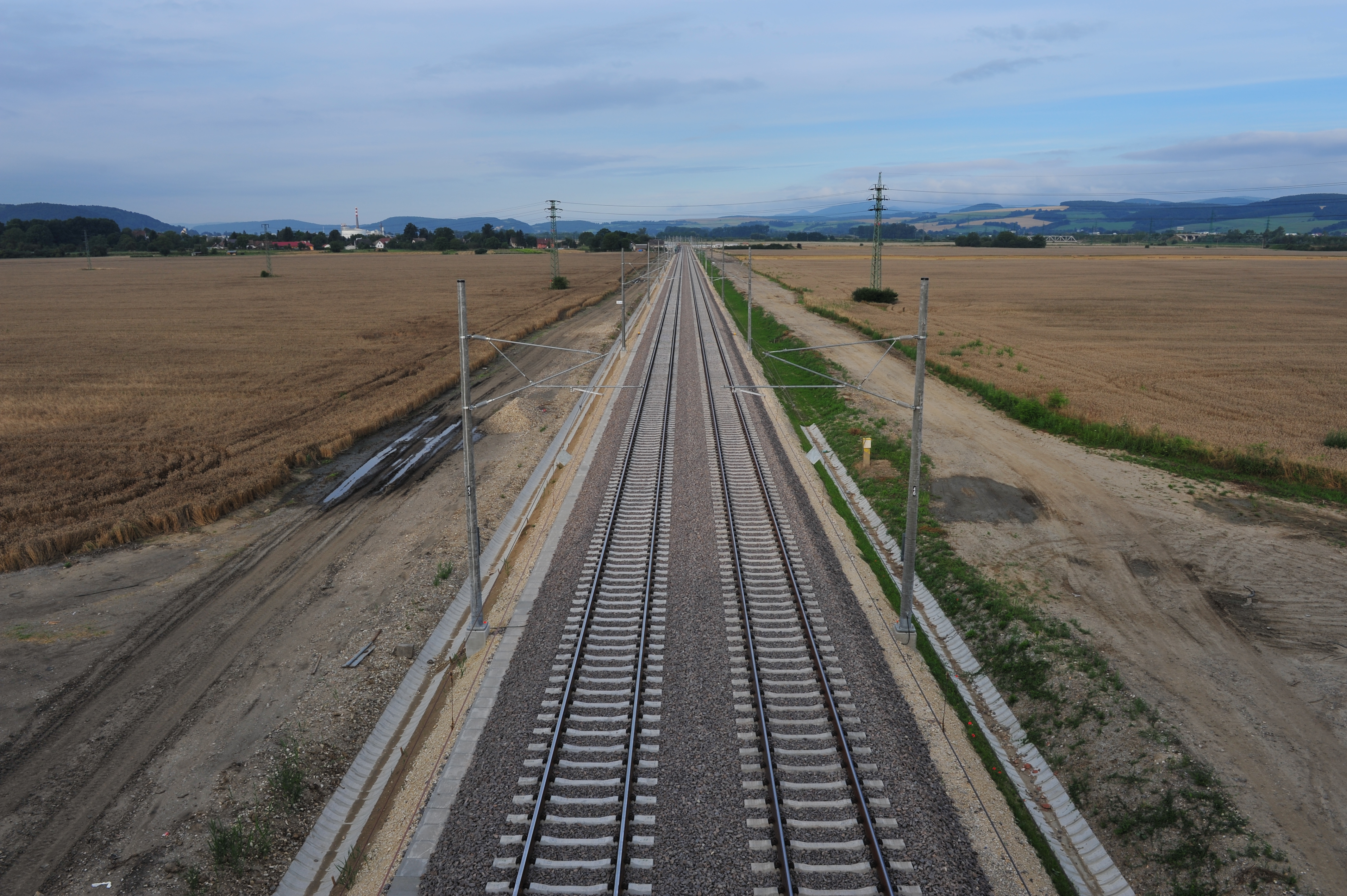 Full speed ahead. Photo: aerial view of two pairs of railway tracks through the fields in vanishing point perspective; power lines and catenary, muddy roads and fields on the side of the tracks, landscape in the background, cloudy evening sky